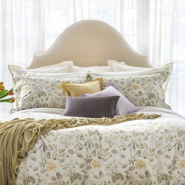 Printed bedding with wildflower design in soft greens, lilac and yellow tones on a soft white background. Celadon coloured Divina velvet throw strewn on bed, and throw pillows in solid purples and yellow at top of bed.